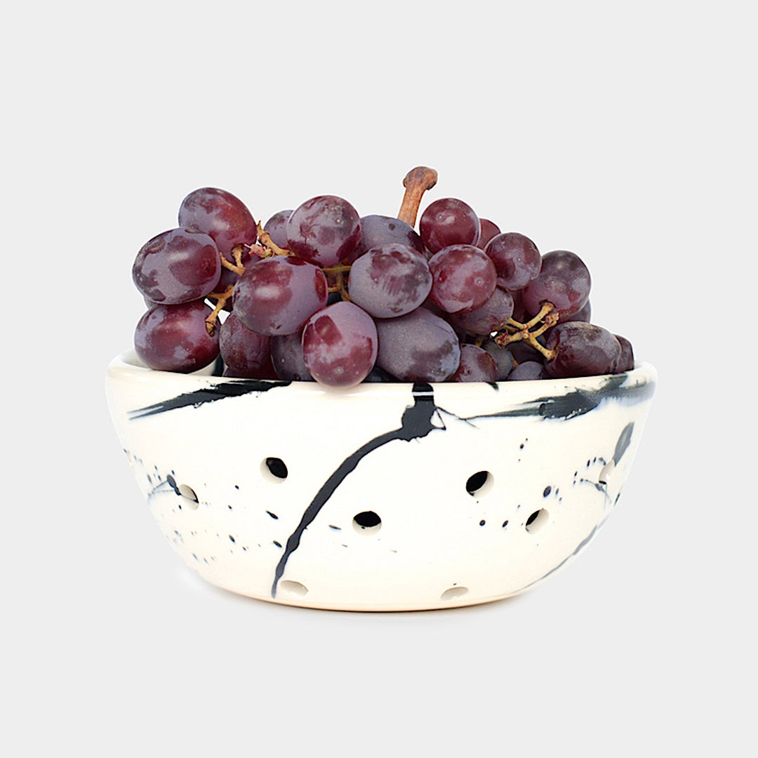 The Earl Home COLANDER is a hand-thrown ceramic bowl with a blue splatter design and circular cutouts, serving as both a dish and dishwasher-safe colander. Photographed on a simple background.