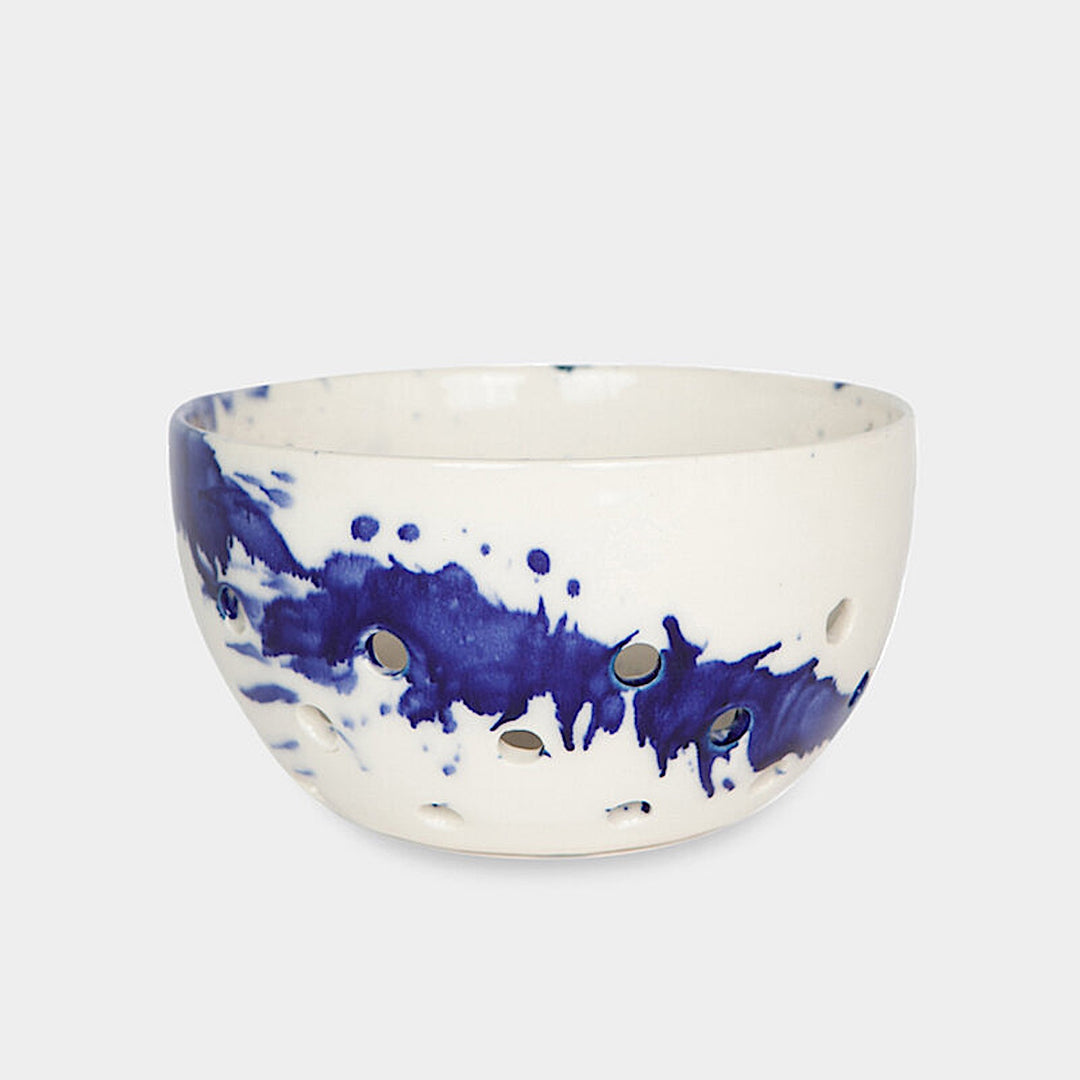 The Earl Home COLANDER is a hand-thrown ceramic bowl with a blue splatter design and circular cutouts, serving as both a dish and dishwasher-safe colander. Photographed on a simple background.