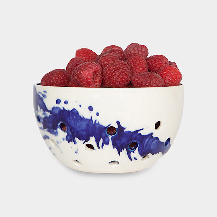 The Earl Home COLANDER is a hand-thrown ceramic bowl with a blue splatter design and circular cutouts, serving as both a dish and dishwasher-safe colander. Photographed on a simple background.