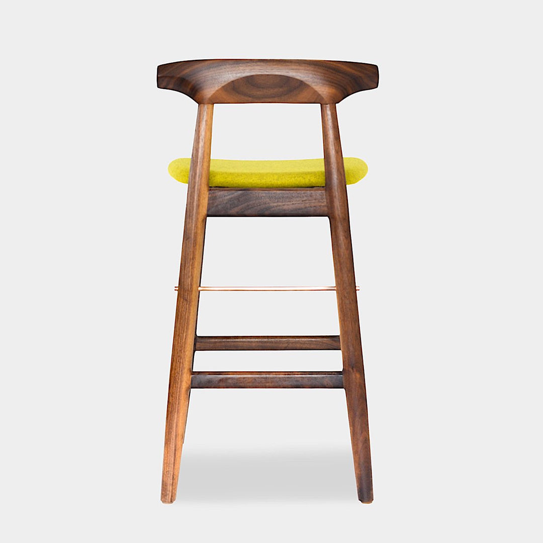 The WOOD High Stool (Bar Height) by Earl Home is a handcrafted wooden bar stool with a light finish, backrest, footrest, and simple design. It offers customizable upholstery to match any decor.