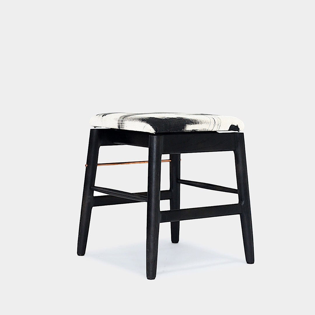 The WOOD Low Stool by Earl Home features a handmade wooden design, a custom upholstered black cushioned seat, and a brass footrest bar, all set against a plain background.