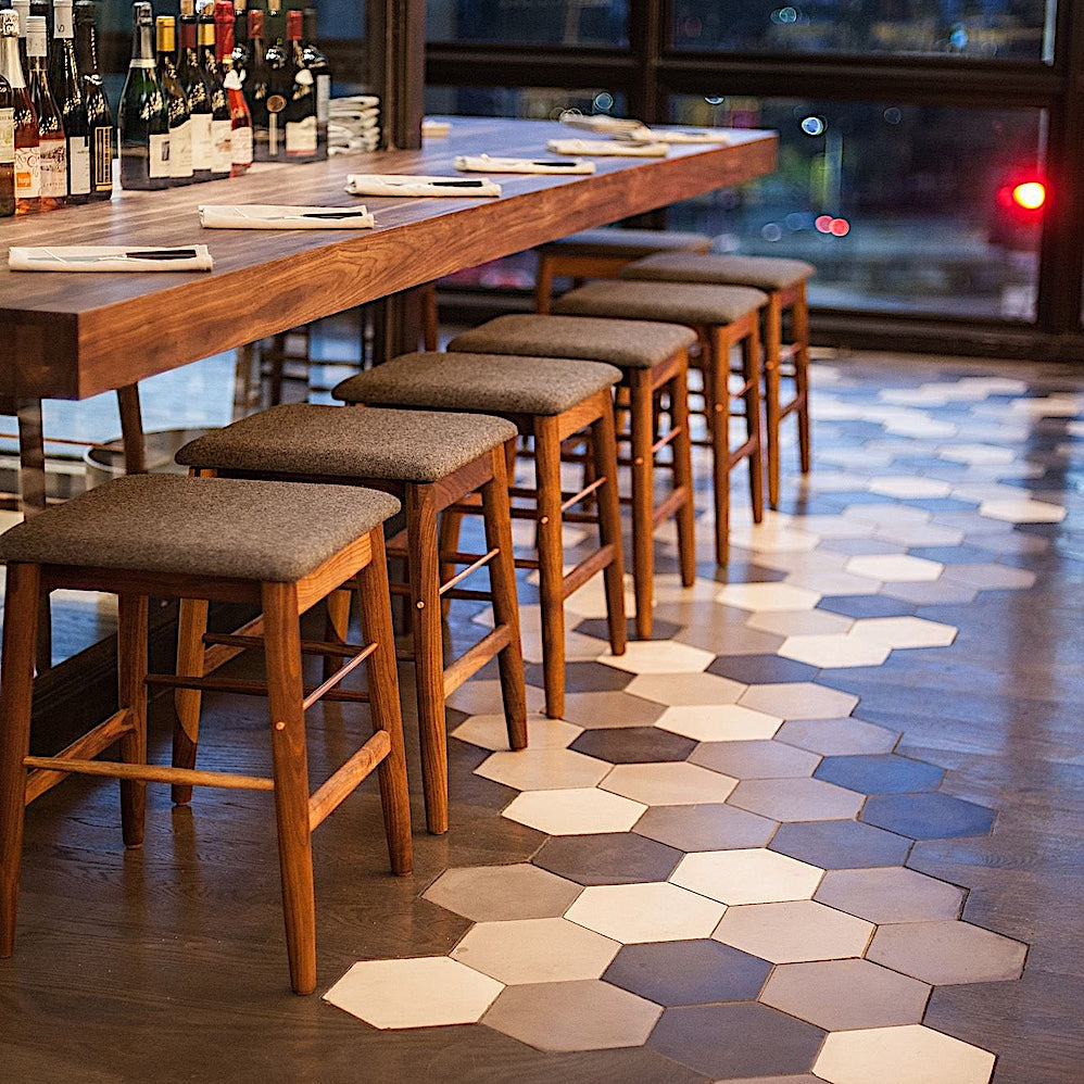 The WOOD Low Stool by Earl Home features a handmade wooden design, a custom upholstered black cushioned seat, and a brass footrest bar, all set against a plain background.