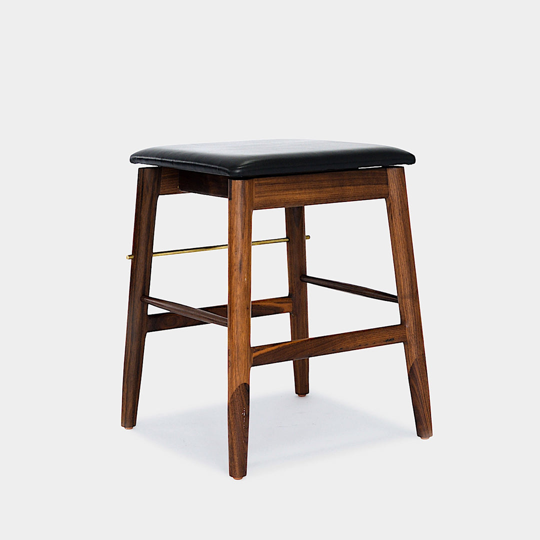 The WOOD Low Stool by Earl Home features a handmade wooden design, a custom upholstered black cushioned seat, and a brass footrest bar, all set against a plain background.