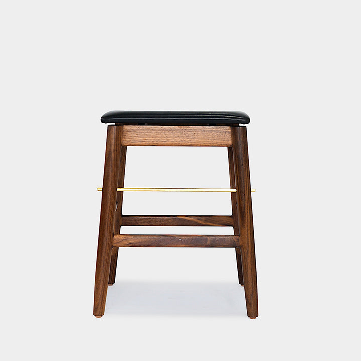 The WOOD Low Stool by Earl Home features a handmade wooden design, a custom upholstered black cushioned seat, and a brass footrest bar, all set against a plain background.