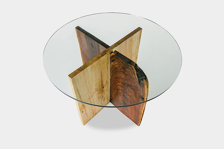 The X Dining Table by Earl Home is a handcrafted, round glass table with intersecting wooden slabs in natural and dark brown tones, showcasing its organic edge and epitomizing wood slab artistry.