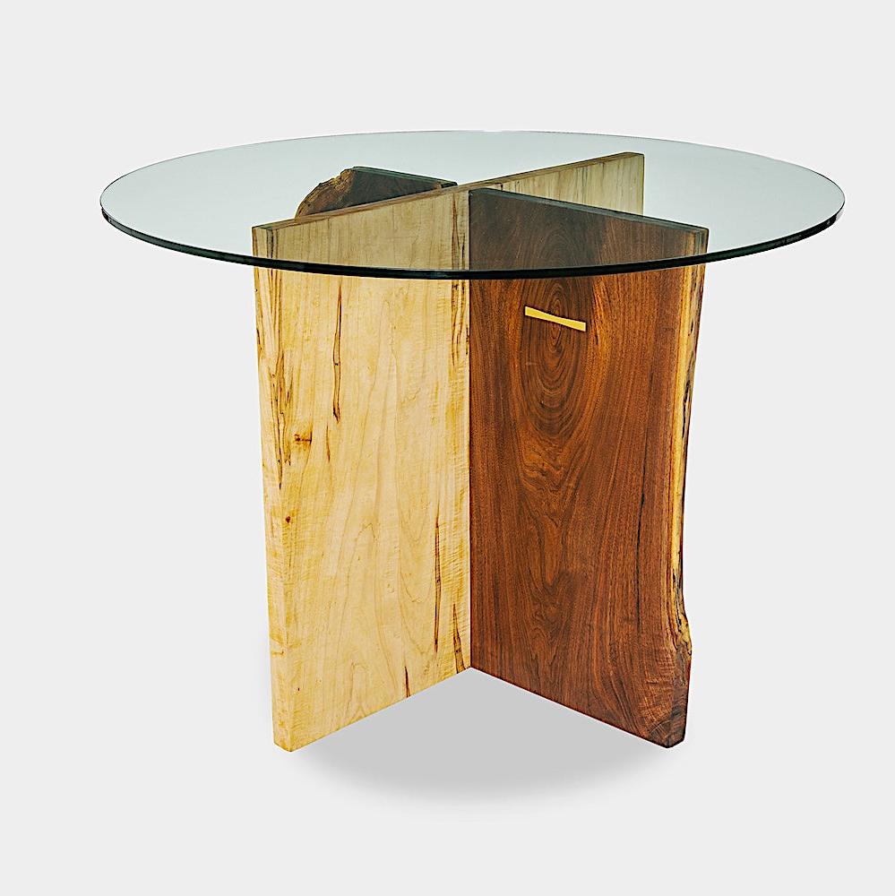 The X Dining Table by Earl Home is a handcrafted, round glass table with intersecting wooden slabs in natural and dark brown tones, showcasing its organic edge and epitomizing wood slab artistry.