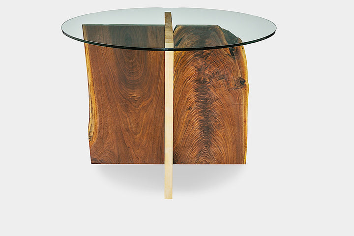 The X Dining Table by Earl Home is a handcrafted, round glass table with intersecting wooden slabs in natural and dark brown tones, showcasing its organic edge and epitomizing wood slab artistry.