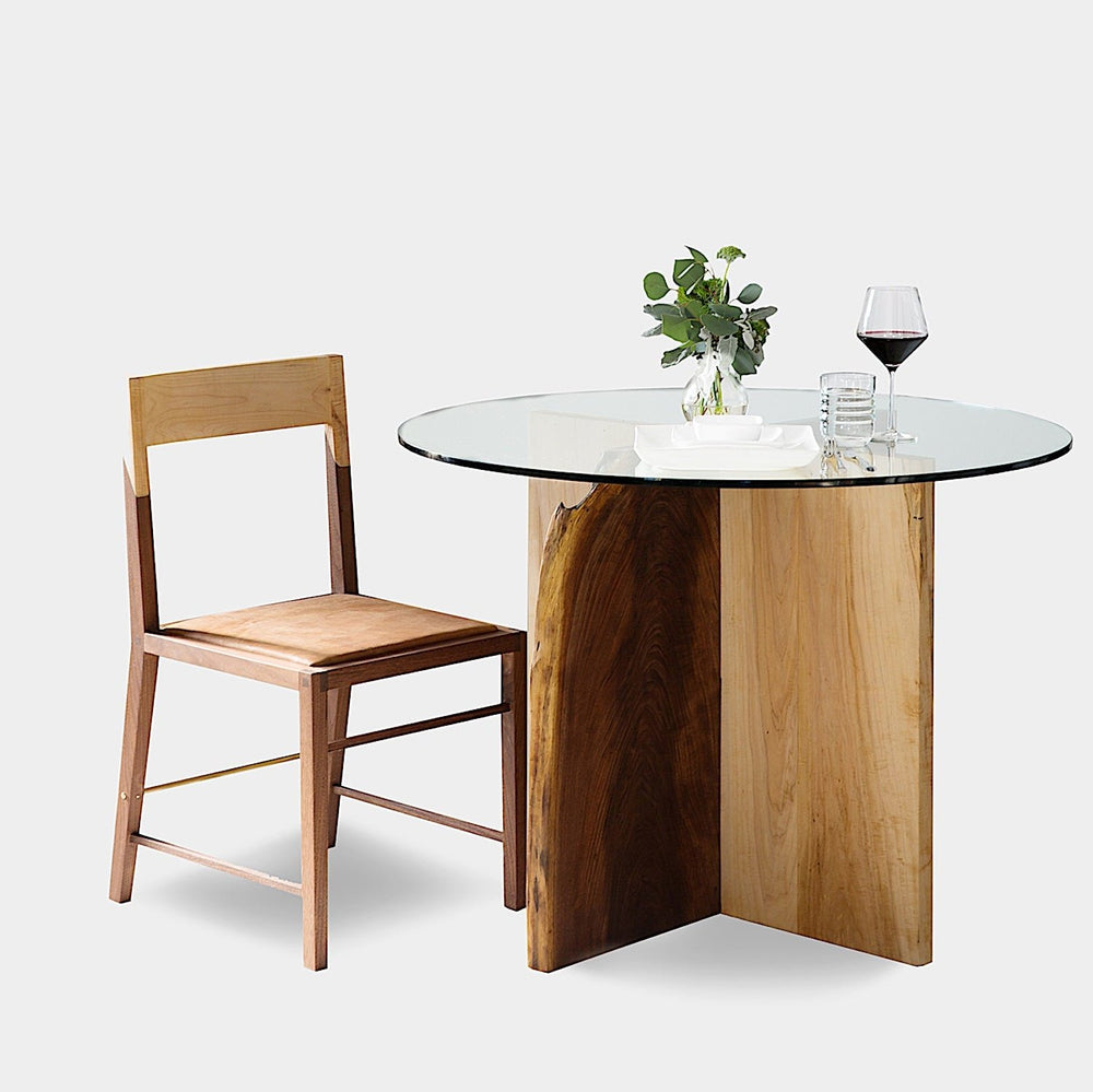 The X Dining Table by Earl Home is a handcrafted, round glass table with intersecting wooden slabs in natural and dark brown tones, showcasing its organic edge and epitomizing wood slab artistry.