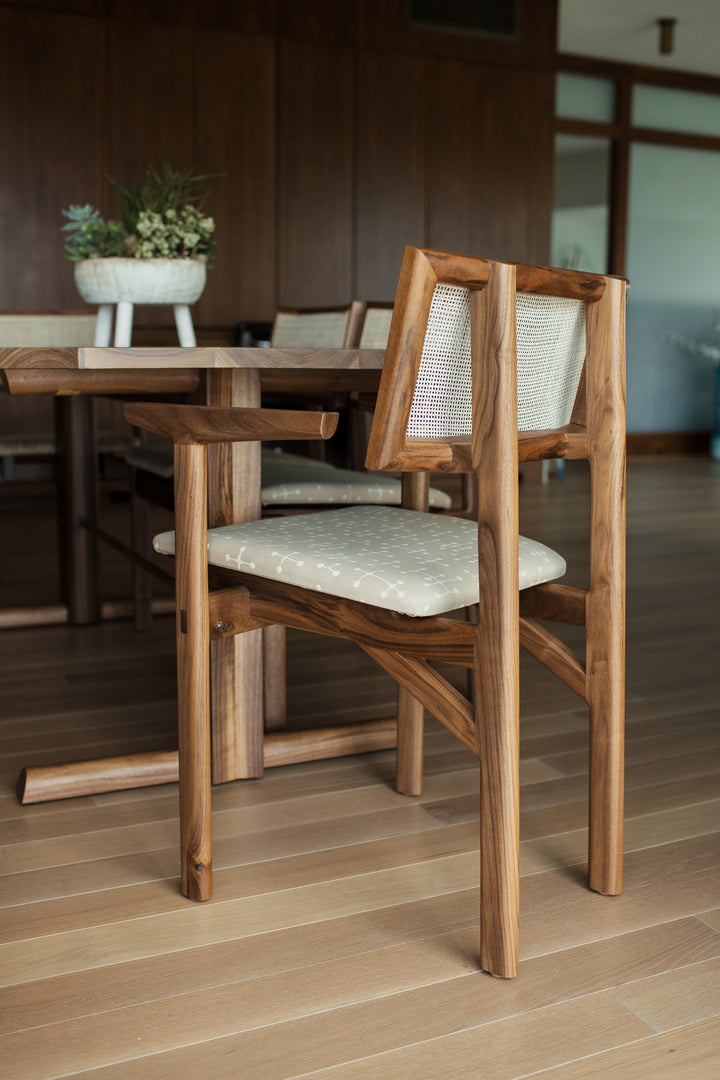 The KUNAI Captain Chair by Earl Home is a solid wood dining chair with armrests, a woven rattan backrest, and a custom upholstered black cushioned seat, all enhanced by intricately hand-shaped legs.