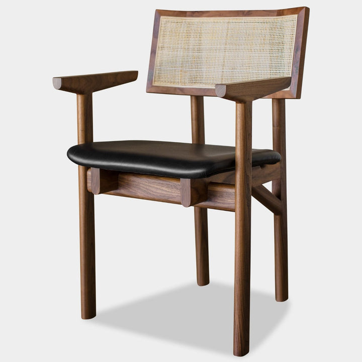 The KUNAI Captain Chair by Earl Home is a solid wood dining chair with armrests, a woven rattan backrest, and a custom upholstered black cushioned seat, all enhanced by intricately hand-shaped legs.