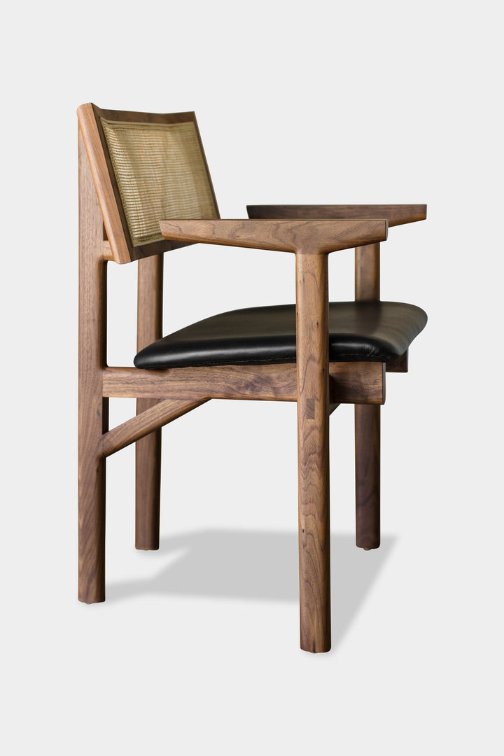 The KUNAI Captain Chair by Earl Home is a solid wood dining chair with armrests, a woven rattan backrest, and a custom upholstered black cushioned seat, all enhanced by intricately hand-shaped legs.