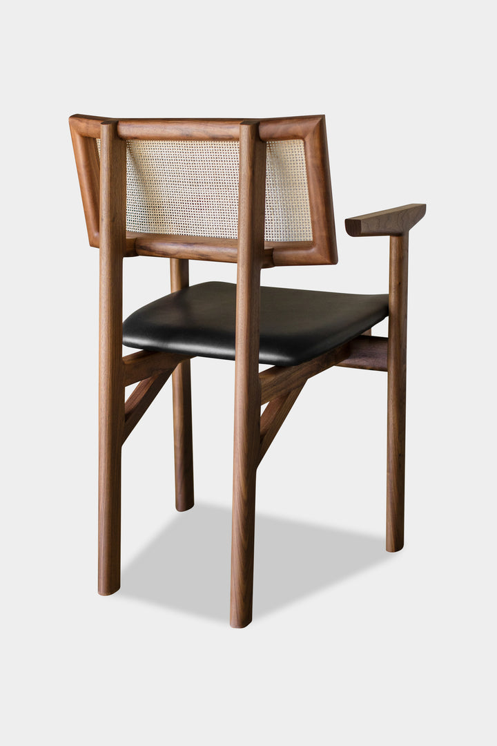 The KUNAI Captain Chair by Earl Home is a solid wood dining chair with armrests, a woven rattan backrest, and a custom upholstered black cushioned seat, all enhanced by intricately hand-shaped legs.