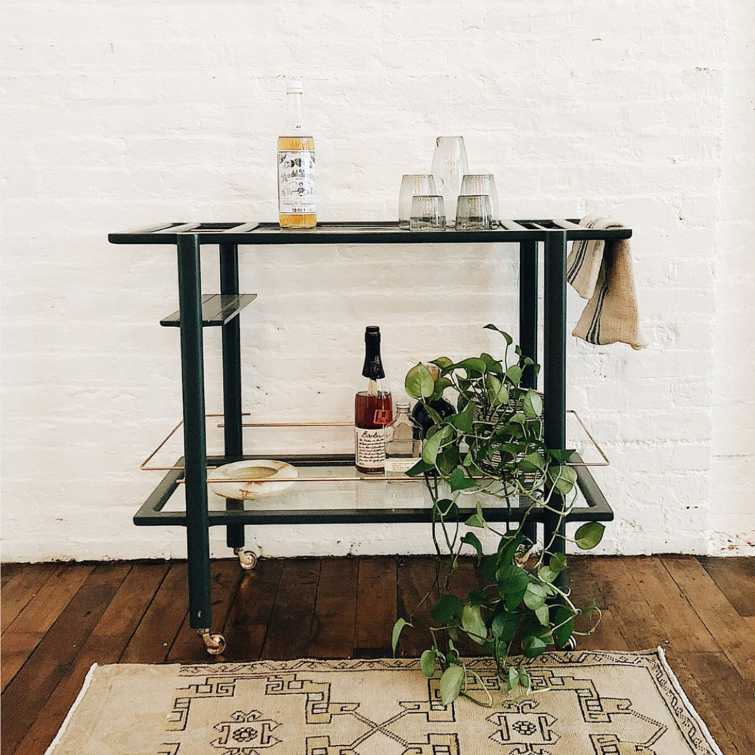 This bespoke BAR Cart from Earl Home is a solid wood bar cart on wheels with exquisite hand-cut joinery, featuring two shelves: a robust tempered glass top and an elegant lower shelf made of clear or smoked tempered glass. This cart comes in walnut, white oak, ebonized oak, or lacquered in any pantone color. Brass, Copper and Aluminum are the metal types offered for detail.