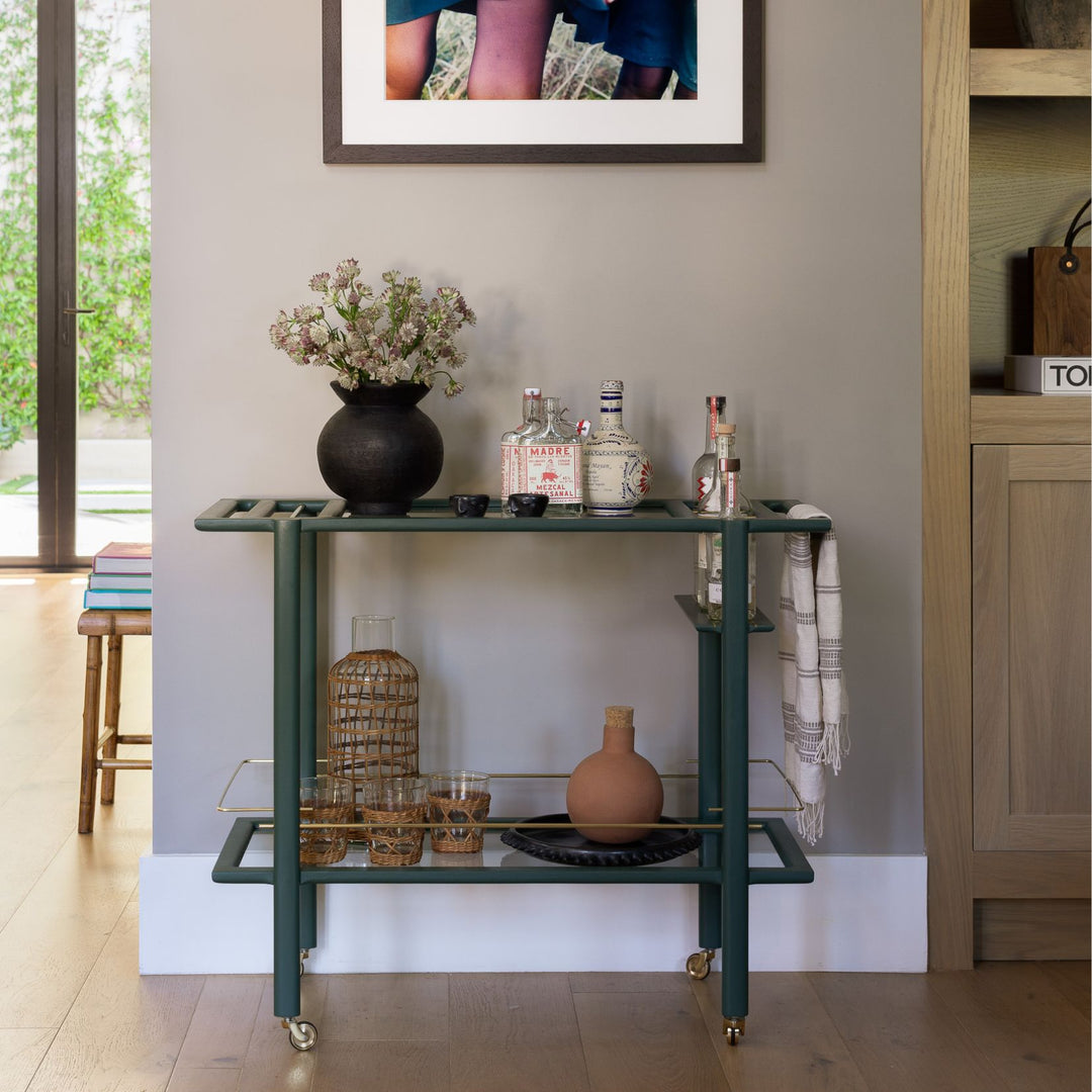 This bespoke BAR Cart from Earl Home is a custom hand made solid wood bar cart on wheels with exquisite hand-cut joinery, featuring two shelves: a robust tempered glass top and an elegant lower shelf made of clear or smoked tempered glass. This cart comes in walnut, white oak, ebonized oak, or lacquered in any pantone color. Brass, Copper and Aluminum are the metal types offered for detail.