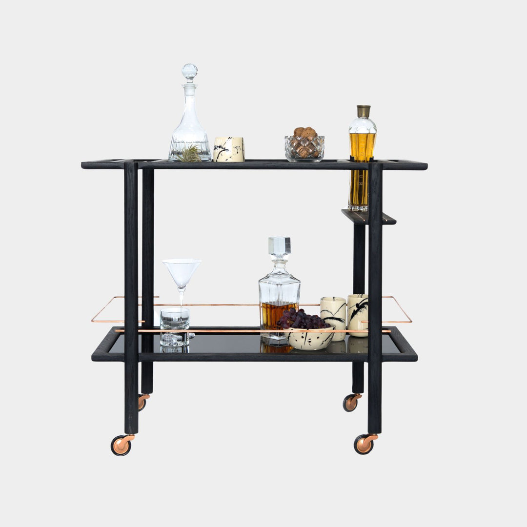 This bespoke BAR Cart from Earl Home is a custom hand made solid wood bar cart on wheels with exquisite hand-cut joinery, featuring two shelves: a robust tempered glass top and an elegant lower shelf made of clear or smoked tempered glass. This cart comes in walnut, white oak, ebonized oak, or lacquered in any pantone color. Brass, Copper and Aluminum are the metal types offered for detail.