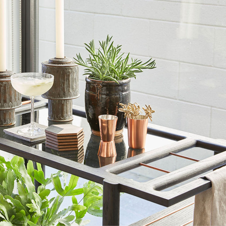 This bespoke BAR Cart from Earl Home is a custom hand made solid wood bar cart on wheels with exquisite hand-cut joinery, featuring two shelves: a robust tempered glass top and an elegant lower shelf made of clear or smoked tempered glass. This cart comes in walnut, white oak, ebonized oak, or lacquered in any pantone color. Brass, Copper and Aluminum are the metal types offered for detail.