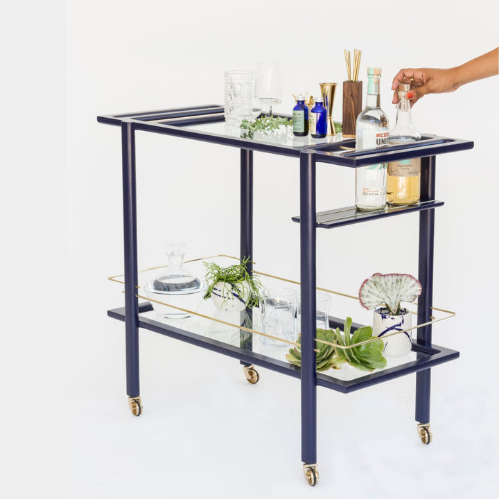 This bespoke BAR Cart from Earl Home is a custom hand made solid wood bar cart on wheels with exquisite hand-cut joinery, featuring two shelves: a robust tempered glass top and an elegant lower shelf made of clear or smoked tempered glass. This cart comes in walnut, white oak, ebonized oak, or lacquered in any pantone color. Brass, Copper and Aluminum are the metal types offered for detail.