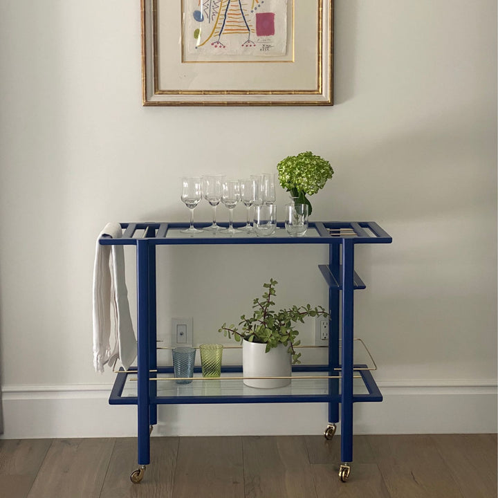 This bespoke BAR Cart from Earl Home is a custom hand made solid wood bar cart on wheels with exquisite hand-cut joinery, featuring two shelves: a robust tempered glass top and an elegant lower shelf made of clear or smoked tempered glass. This cart comes in walnut, white oak, ebonized oak, or lacquered in any pantone color. Brass, Copper and Aluminum are the metal types offered for detail.