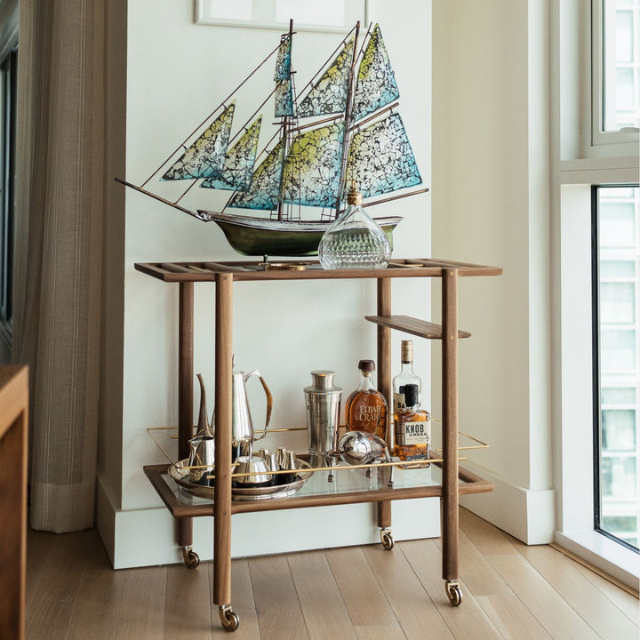 This bespoke BAR Cart from Earl Home is a custom hand made solid wood bar cart on wheels with exquisite hand-cut joinery, featuring two shelves: a robust tempered glass top and an elegant lower shelf made of clear or smoked tempered glass. This cart comes in walnut, white oak, ebonized oak, or lacquered in any pantone color. Brass, Copper and Aluminum are the metal types offered for detail.