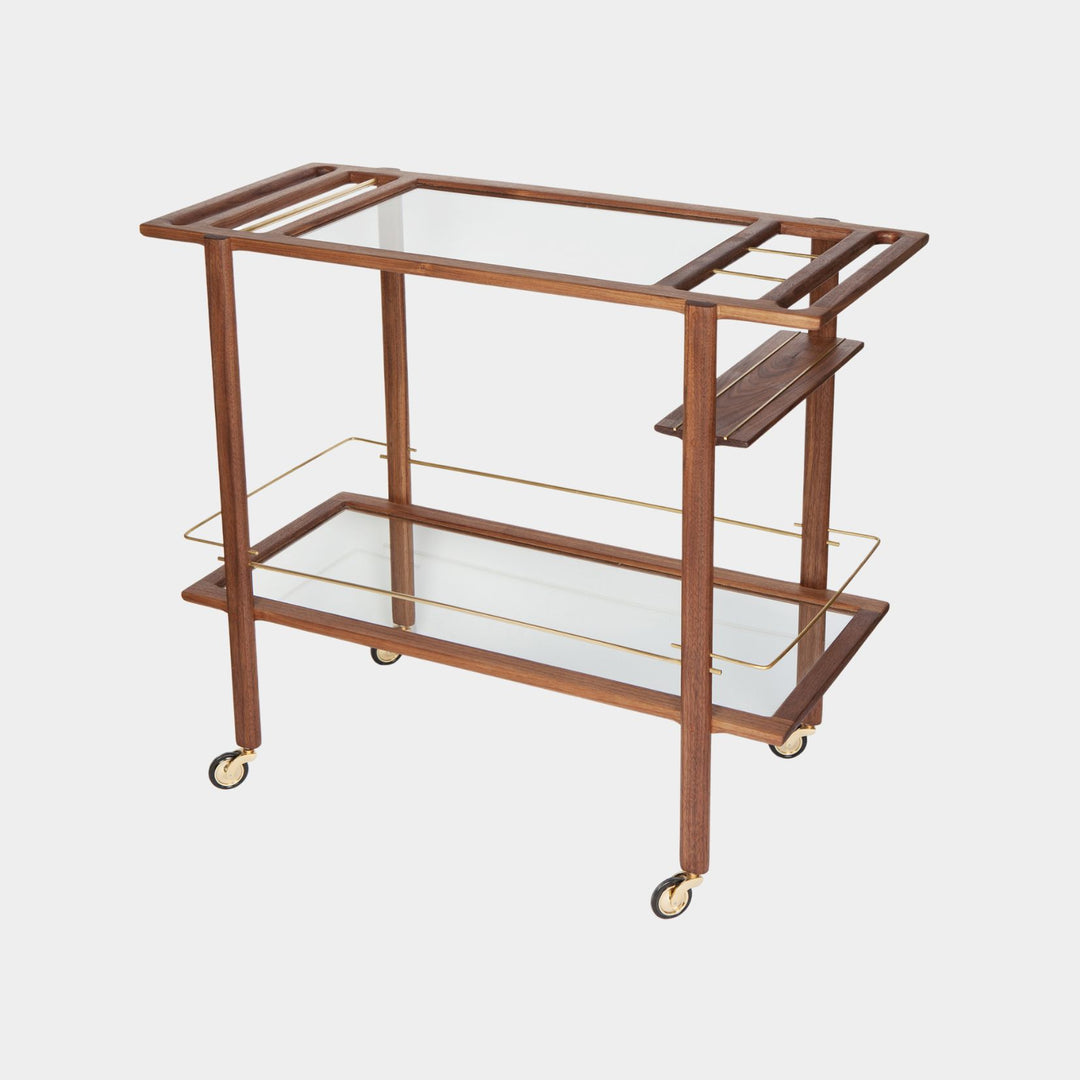 This bespoke BAR Cart from Earl Home is a custom hand made solid wood bar cart on wheels with exquisite hand-cut joinery, featuring two shelves: a robust tempered glass top and an elegant lower shelf made of clear or smoked tempered glass. This cart comes in walnut, white oak, ebonized oak, or lacquered in any pantone color. Brass, Copper and Aluminum are the metal types offered for detail.