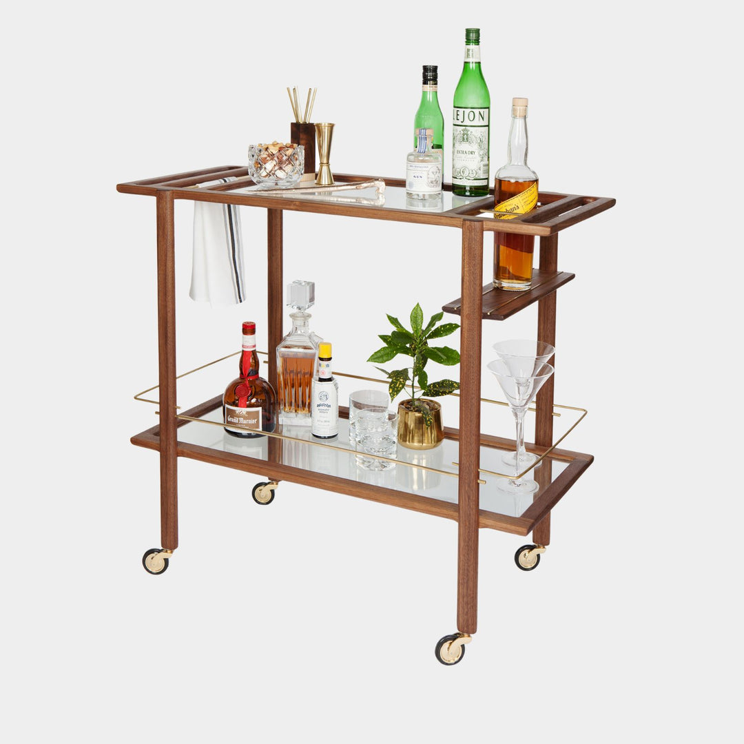 This bespoke BAR Cart from Earl Home is a custom hand made solid wood bar cart on wheels with exquisite hand-cut joinery, featuring two shelves: a robust tempered glass top and an elegant lower shelf made of clear or smoked tempered glass. This cart comes in walnut, white oak, ebonized oak, or lacquered in any pantone color. Brass, Copper and Aluminum are the metal types offered for detail.