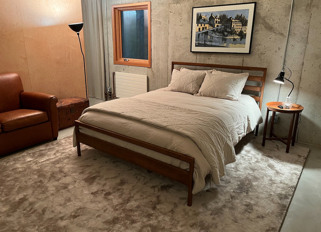 A cozy bedroom with a wooden bed, beige comforter, a brown armchair, wooden side table with a lamp, and a picture on a concrete wall.