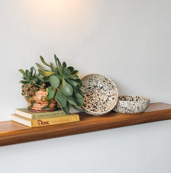 Wooden shelf with plants and books, accompanied by wooden stools against a white wall.