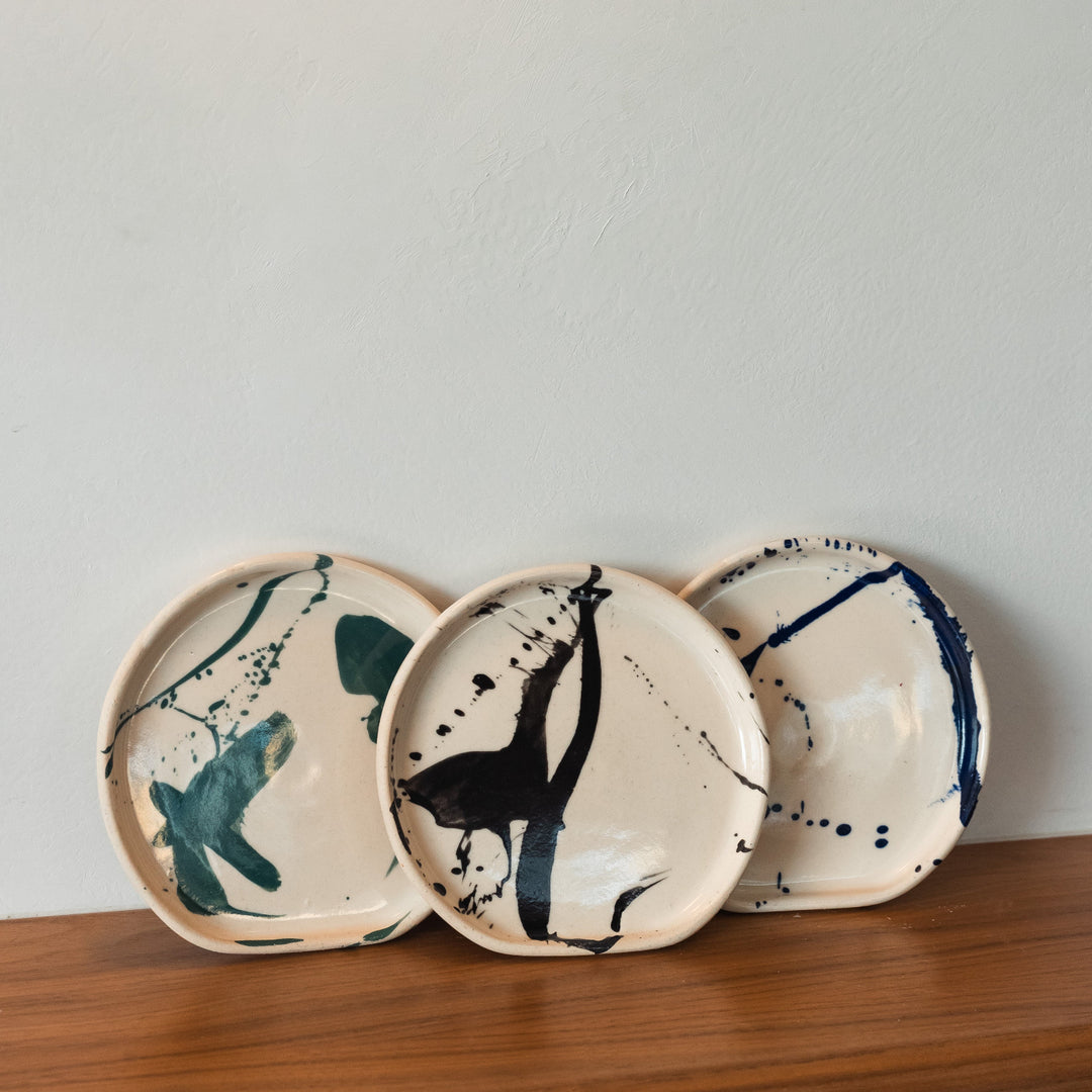 Three ceramic plates with abstract designs on a wooden shelf against a light gray wall.