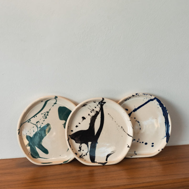 Three ceramic plates with abstract designs on a wooden shelf against a light gray wall.