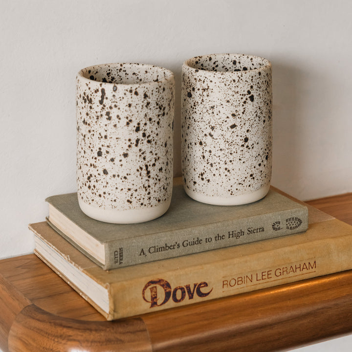 Two speckled ceramic cups on a stack of books with a wooden surface and light background