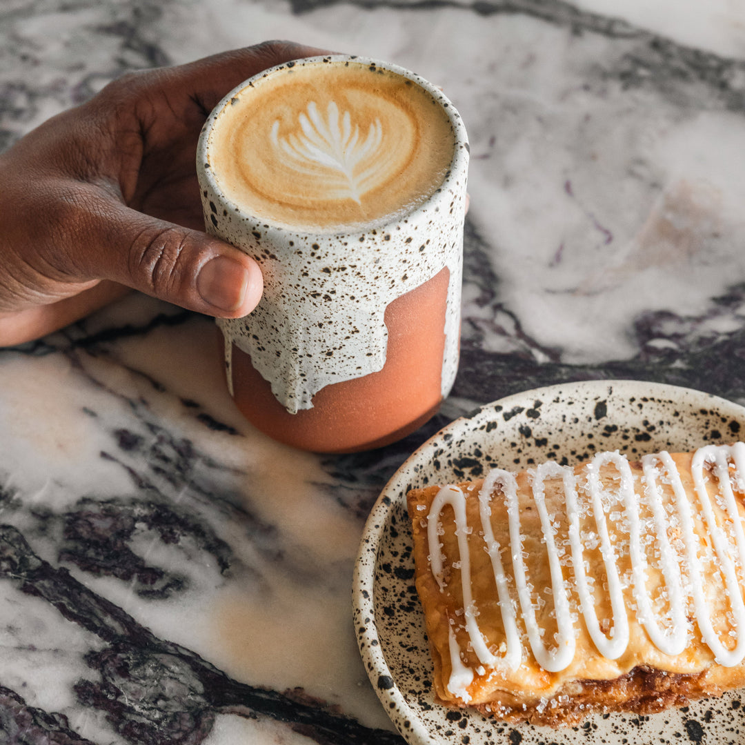 Hand holding a speckled ceramic cup with a latte art design, next to a plate of dessert on a marble surface.