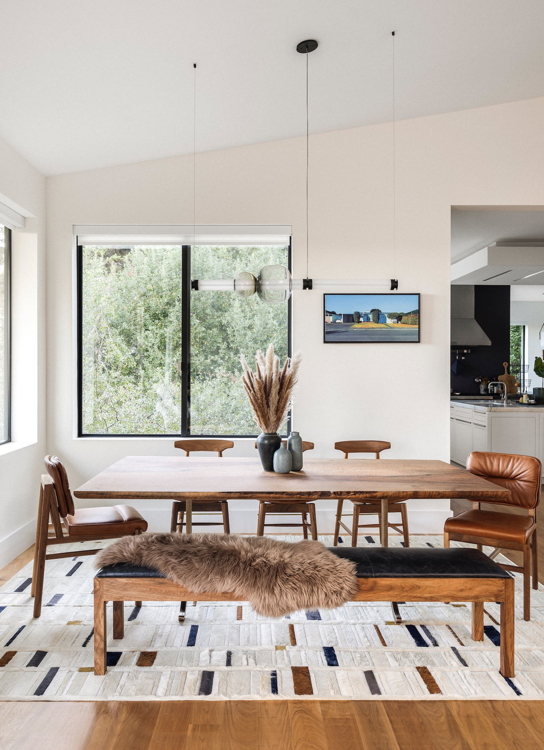 Modern dining room with wooden table, six chairs, bench with fur throw, and two vases with pampas grass. Large windows display a green outdoor view.