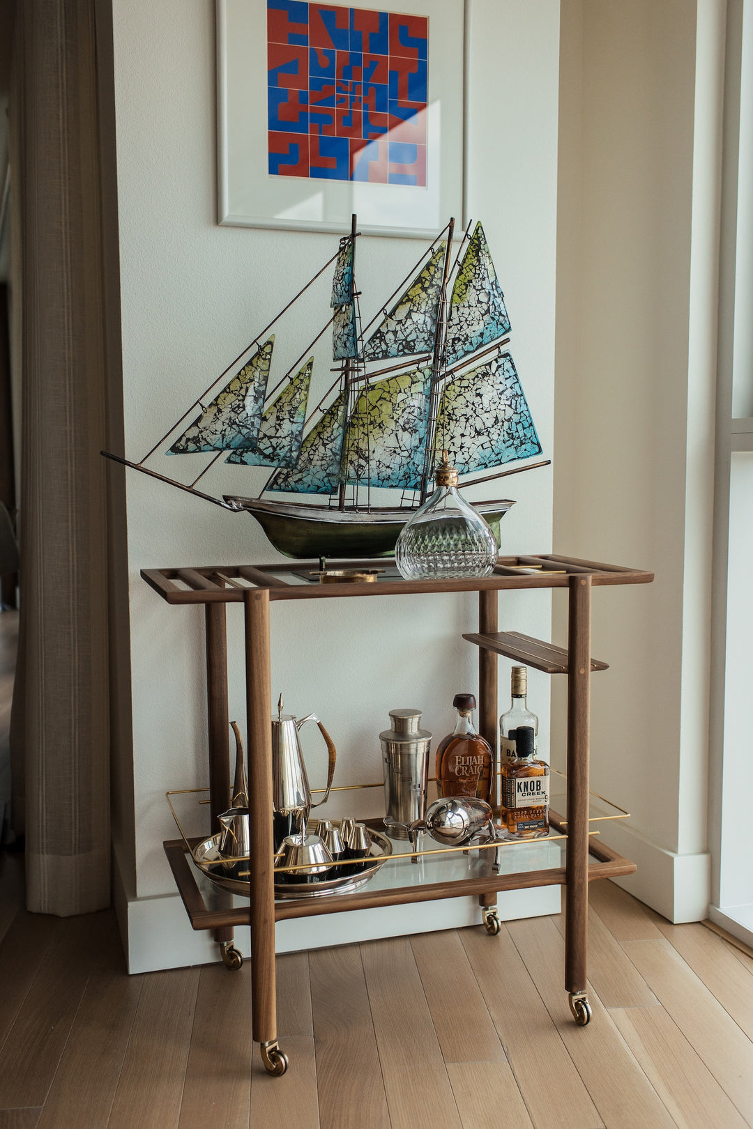 A wooden bar cart with a glass shelf holds glassware, a decanter, a bottle of whiskey, and a model sailboat. A modern art piece hangs on the wall above it.