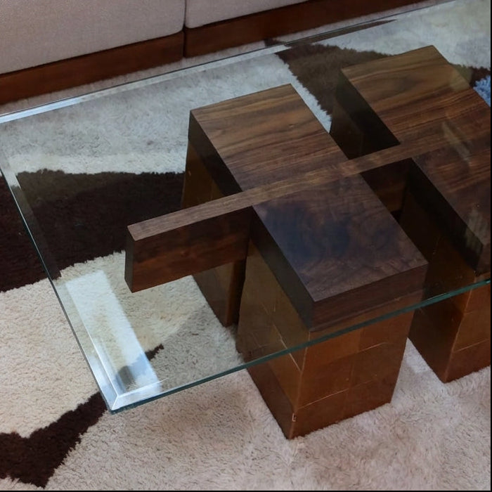 A modern glass-top coffee table with dark wood block bases, placed on a patterned brown and cream rug in front of a light beige sofa.