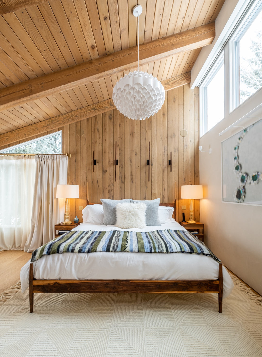 A cozy bedroom with a wooden ceiling and walls, featuring a bed with striped bedding, two side tables with lamps, and a large pendant light above. Natural light fills the space.