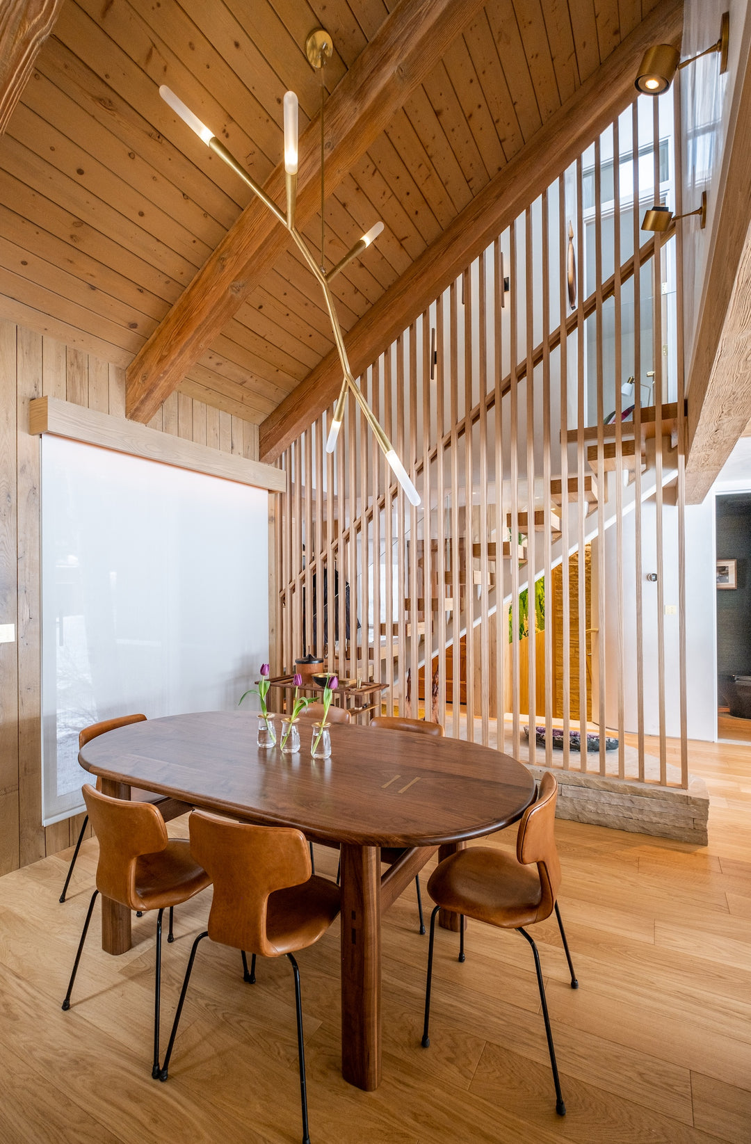 A modern dining area with a wooden table, six chairs, and a stylish ceiling light. The background features a wooden staircase with vertical slats.