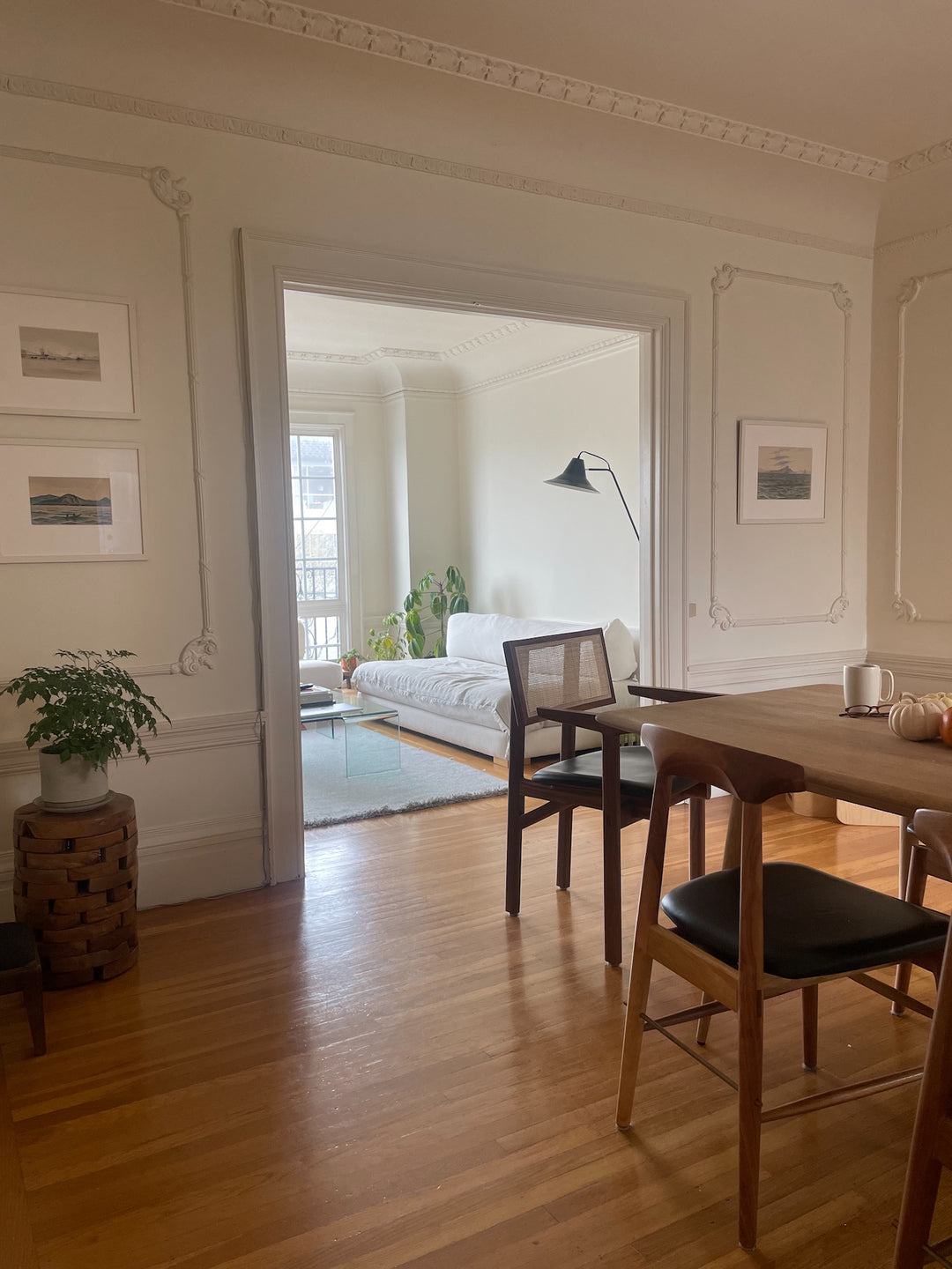 A bright room with a wooden floor, featuring a dining table and chairs in the foreground and a sofa, floor lamp, and plants in the background. Walls are adorned with framed artwork.