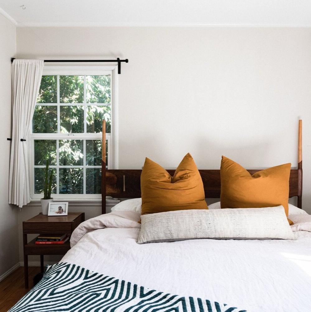 Bedroom with a window, two mustard pillows on a wooden bed, a long cushion, and a bedside table with books and a framed photo.