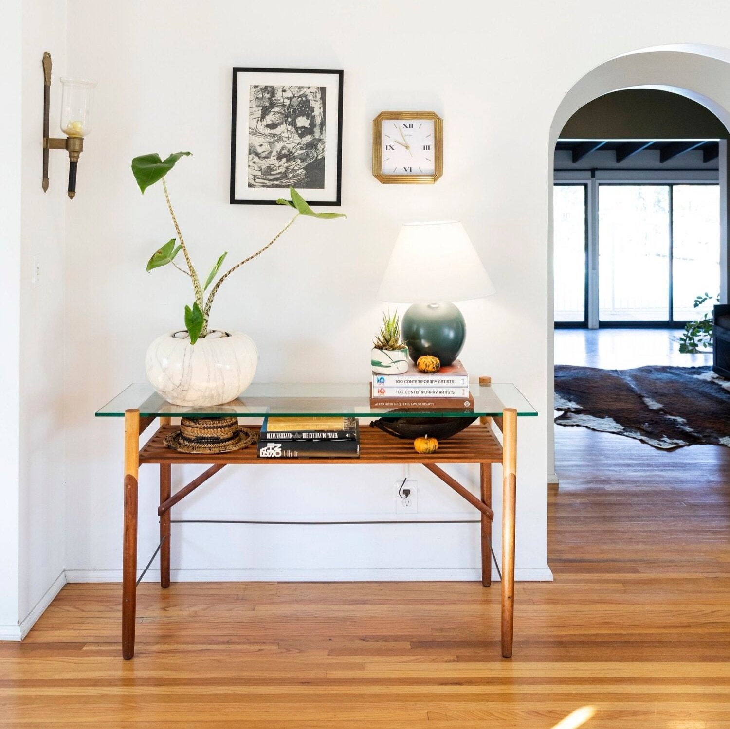 Glass-top table in a hallway with a potted plant, books, a lamp, and a clock. Wall art and a sconce are mounted above. Archway leads to a bright room with a rug.