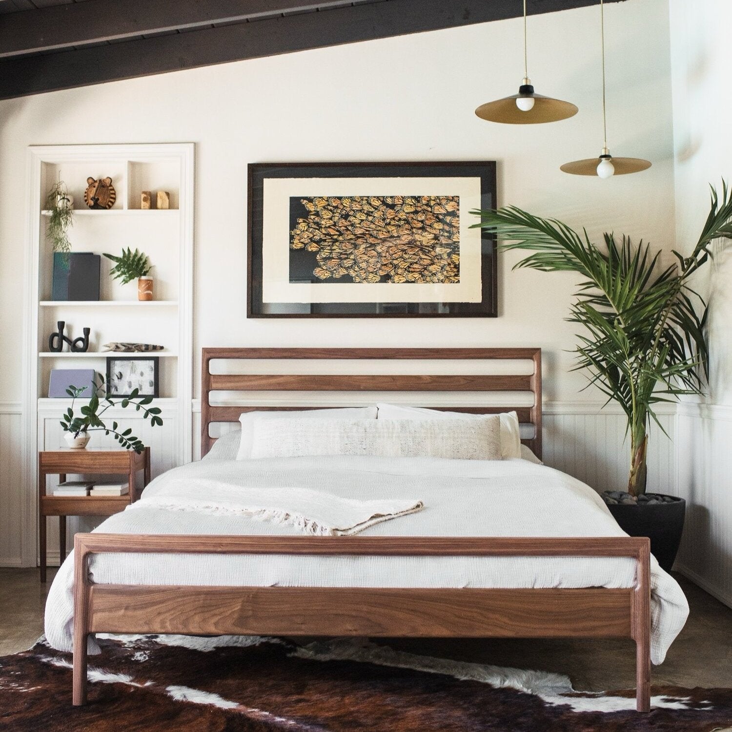 Bedroom with a wooden bedframe, white bedding, two hanging lights, plants, and framed artwork on the wall. Shelving with books and decor is on the left.