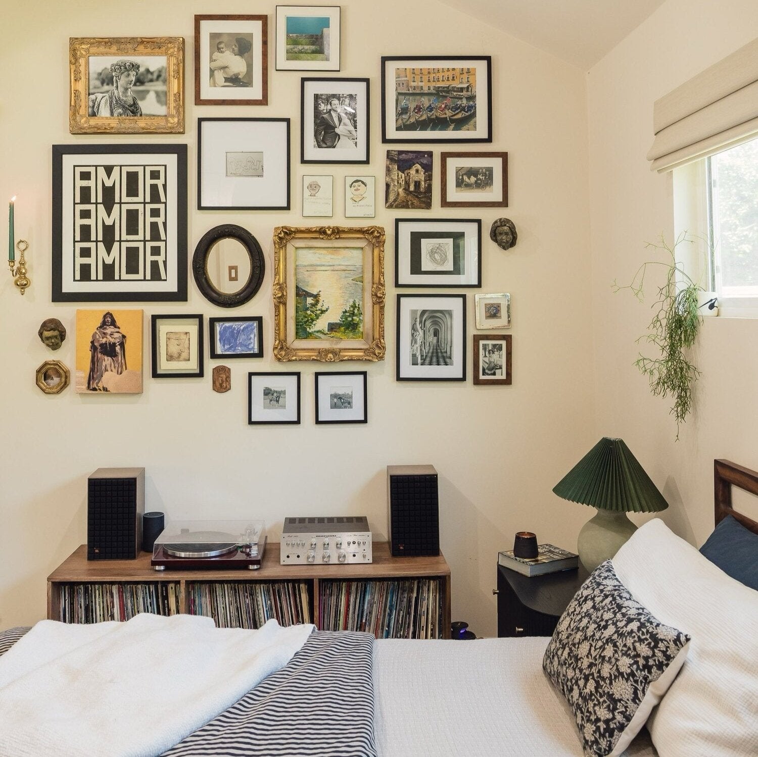 A bedroom with a gallery wall of various framed artworks above a vinyl record setup. A bed with white and patterned bedding is in the foreground, and a green lamp is on a wooden side table.