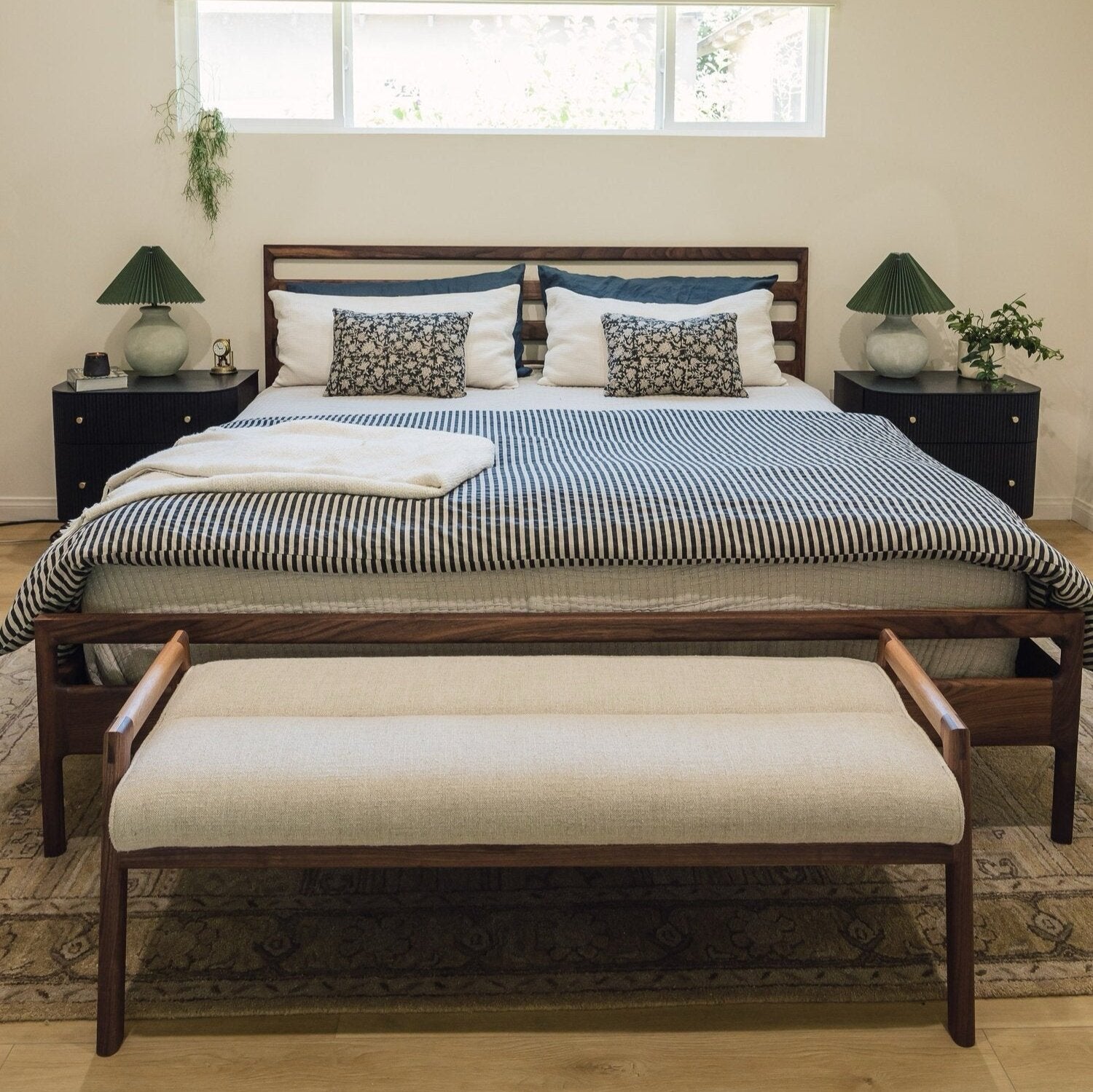 A neatly made bed with a striped duvet and four pillows, flanked by two bedside tables, each with a lamp, under a set of windows. A cushioned bench sits at the foot of the bed.