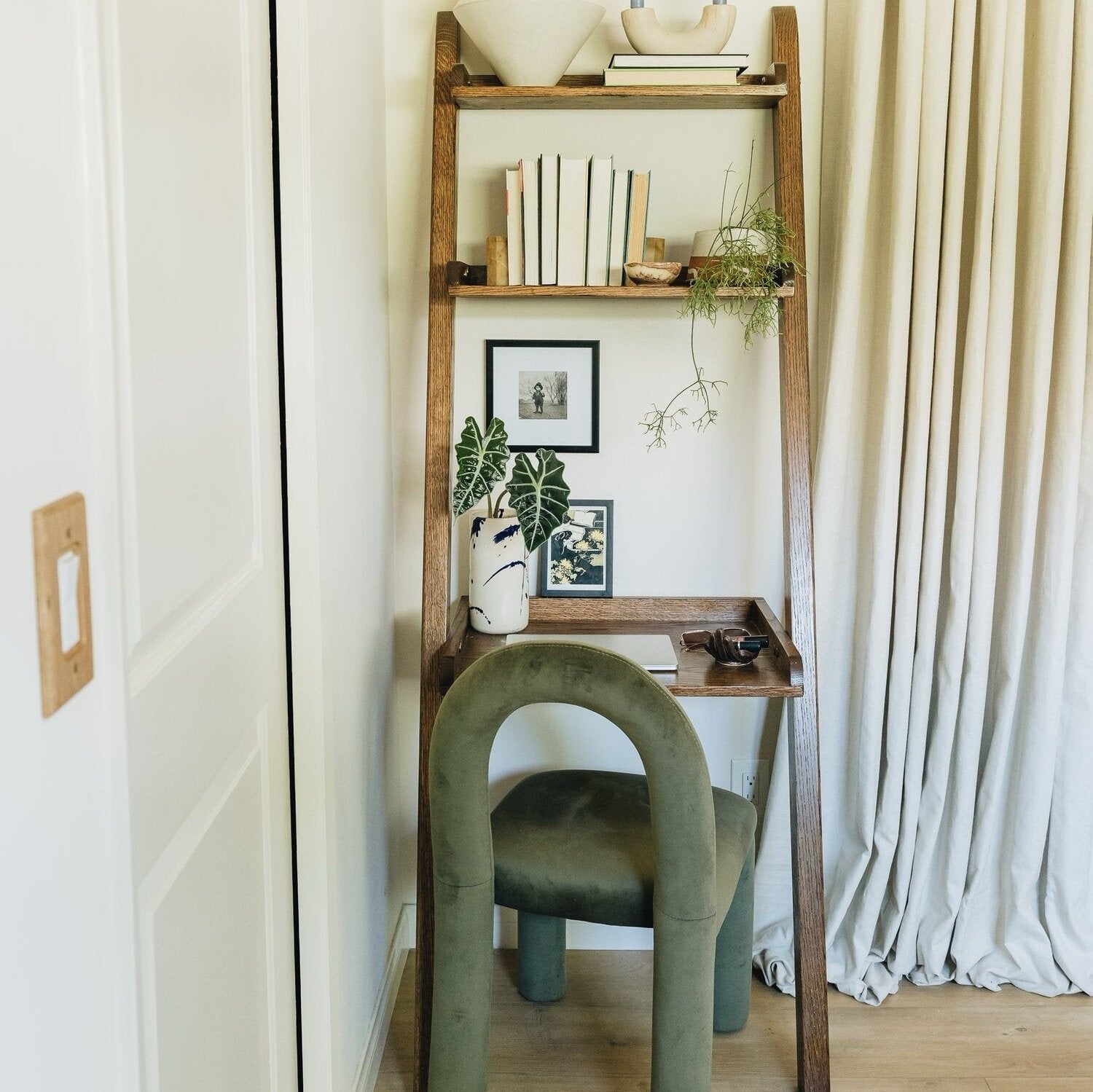 Ladder shelf with books, plants, and framed photos beside an arch-shaped green chair in a corner room with a curtain.