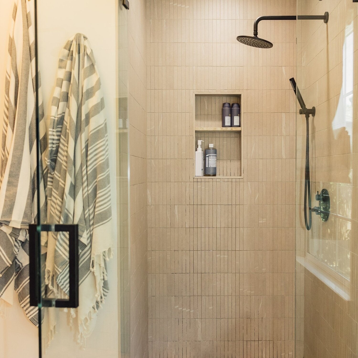 Modern shower with beige tile, overhead and handheld showerheads. Built-in shelf holds toiletries. Stripe-patterned towels hang on the glass door.