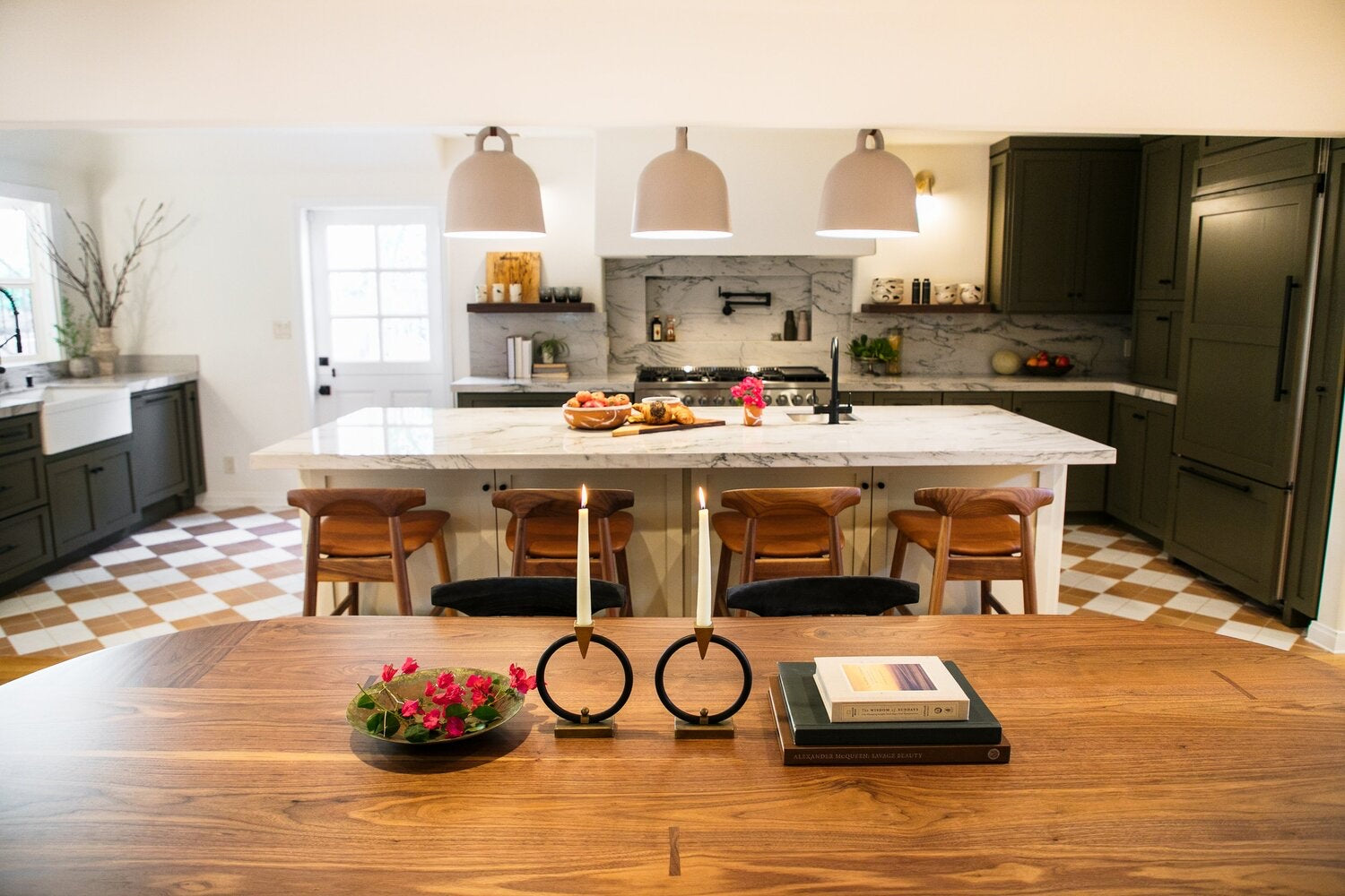 A modern kitchen with a large island, wooden chairs, pendant lights, and a marble backsplash. A dining table with books, candlesticks, and a small flower arrangement is in the foreground.