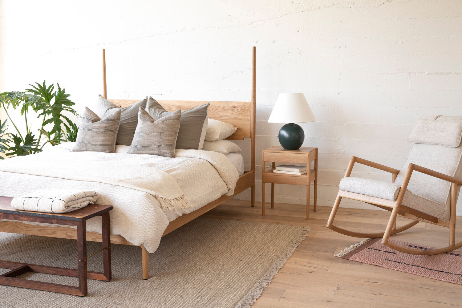 A minimalist bedroom with a wooden bed, gray pillows, a nightstand with a lamp, a rocking chair, and a bench. A plant decorates the corner, and rugs cover the light wood floor.