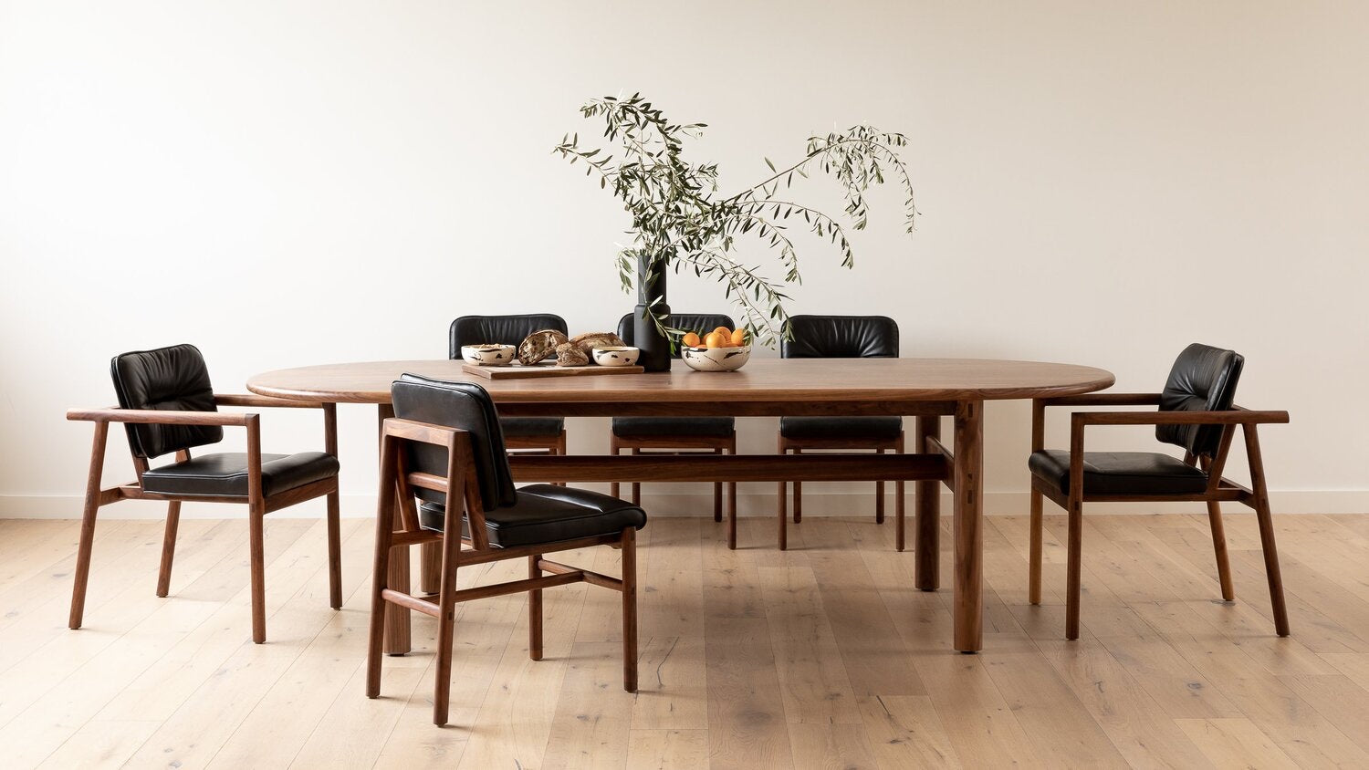 A wooden dining table with six black cushioned chairs is arranged in a room. A vase with branches and bowls of fruit are placed on the table. The floor is wooden, and the walls are plain white.