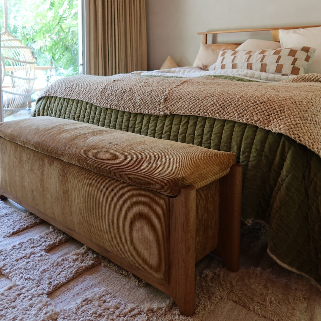 A cozy bedroom scene featuring a bed dressed in textured beige and olive green bedding, a matching upholstered bench at the foot, a wood-paneled ceiling, and a large window with beige curtains.