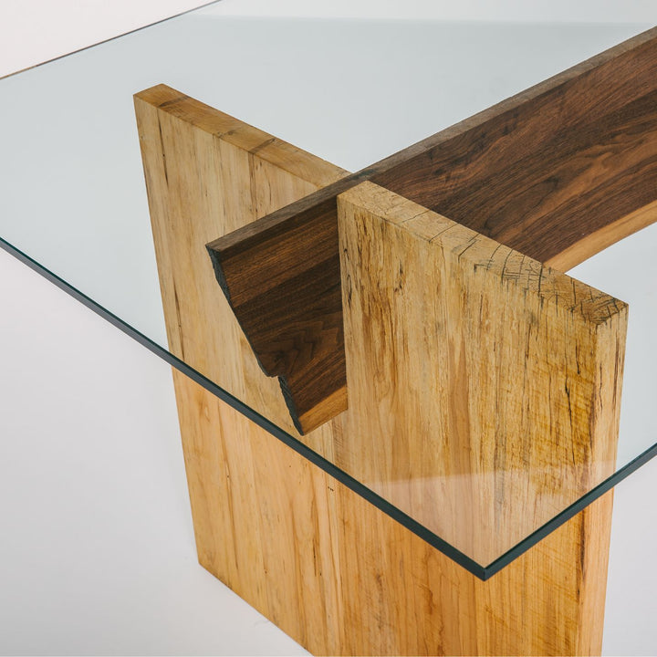 The LUXE Dining Table by Earl Home features a polished glass top and a wooden base with natural wood grain and geometric lines. Solid maple uprights with a walnut spine.