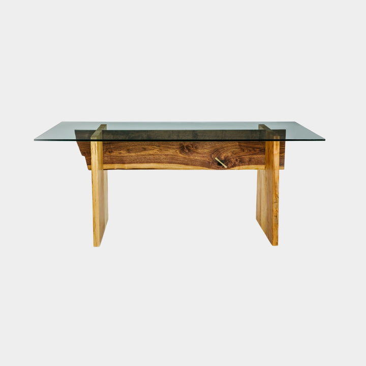 The LUXE Dining Table by Earl Home features a polished glass top and a wooden base with natural wood grain and geometric lines. Solid maple uprights with a walnut spine.